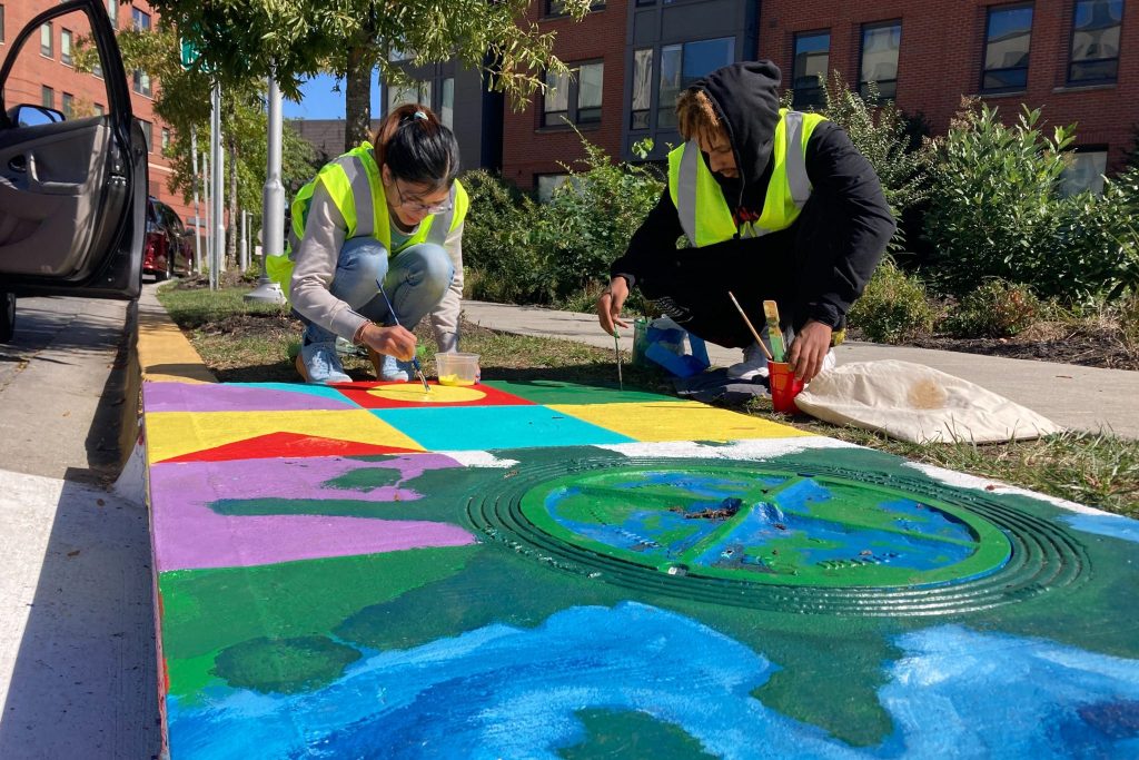 Carla Duran and Beniyam Berhanu, UMD student artists, paint a storm drain along Guilford Drive on Oct. 10, 2022.