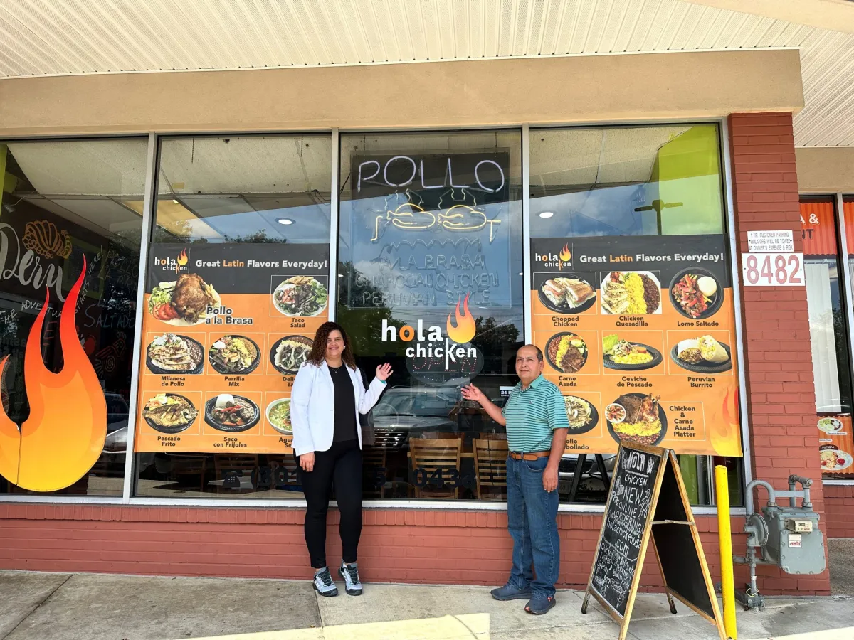 Two people stand in front of a restaurant called Hola Chicken